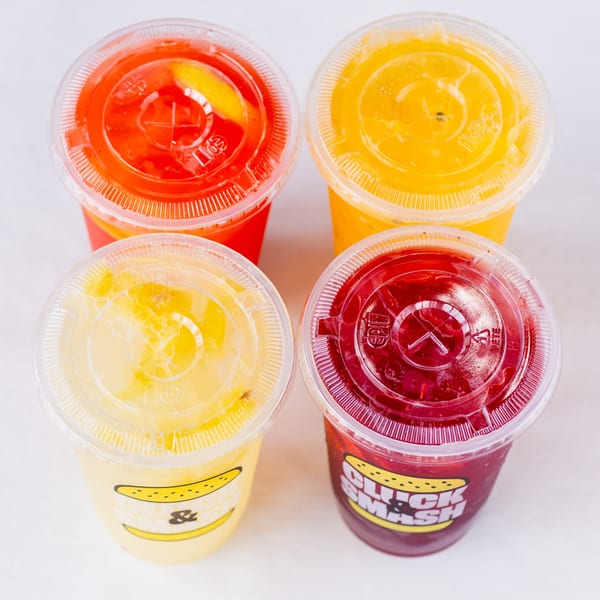 Four colorful Cluck & Smash lemonades in plastic cups with lids, two yellow, one orange, one red.
