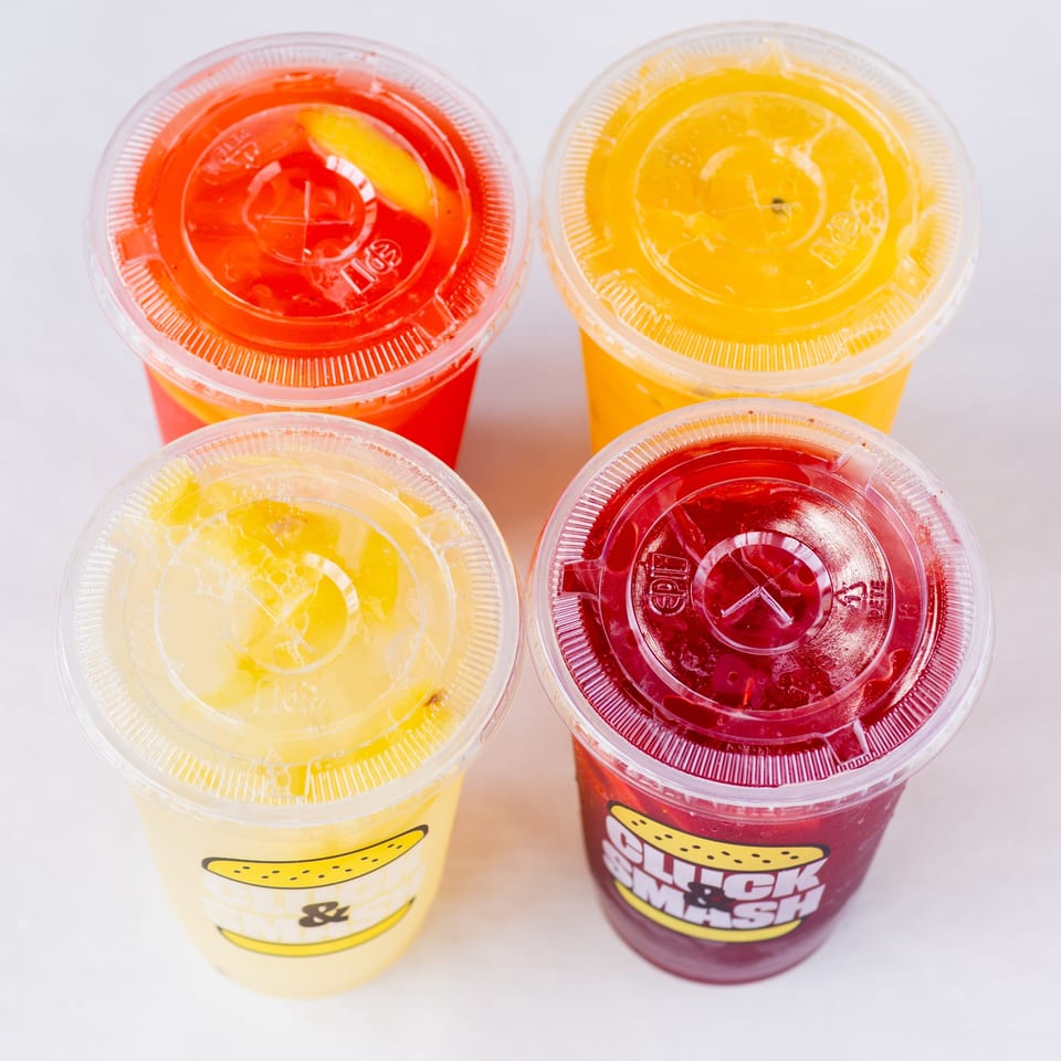 Four colorful Cluck & Smash lemonades in plastic cups with lids, two yellow, one orange, one red.