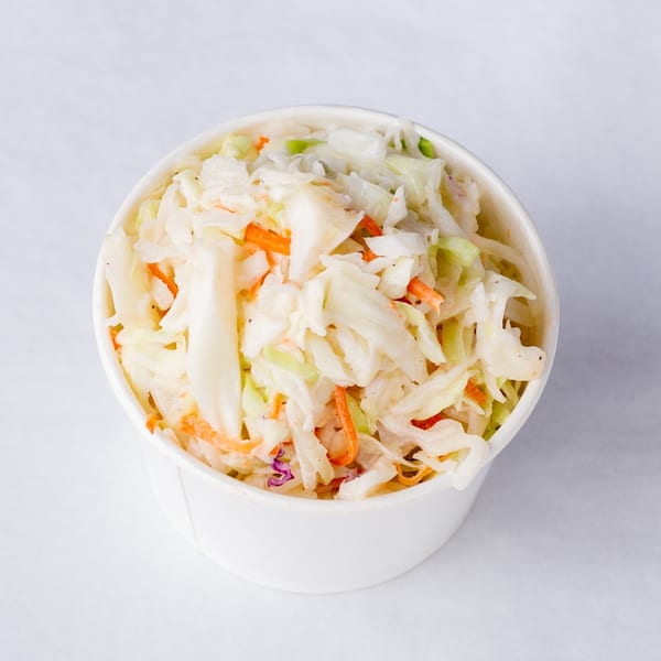 A white paper cup filled with creamy coleslaw, featuring shredded cabbage and carrots.