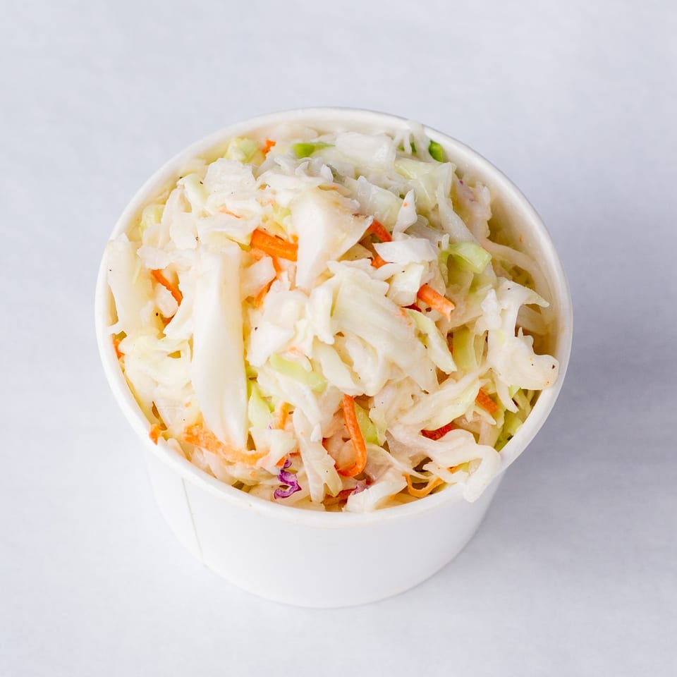 A white paper cup filled with creamy coleslaw, featuring shredded cabbage and carrots.