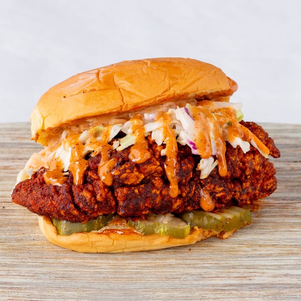 Cluck & Smash fried chicken sandwich with coleslaw, pickles, and orange sauce on a bun.