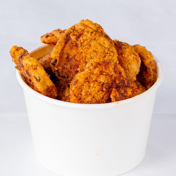 A white bucket filled with crispy, golden-brown fried chicken pieces, seasoned with dark spices.